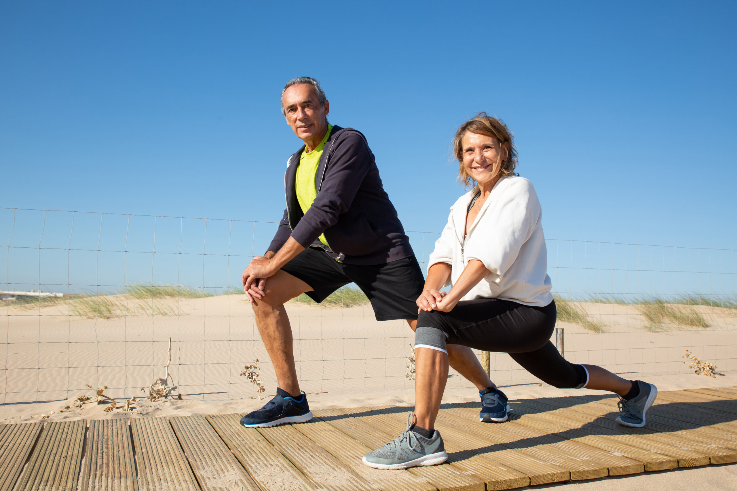 Portrait of happy elderly couple doing workout together. Caucasian man and woman in sportswear half sitting stretching legs smiling looking at camera. Free time for sport activity when retired concept