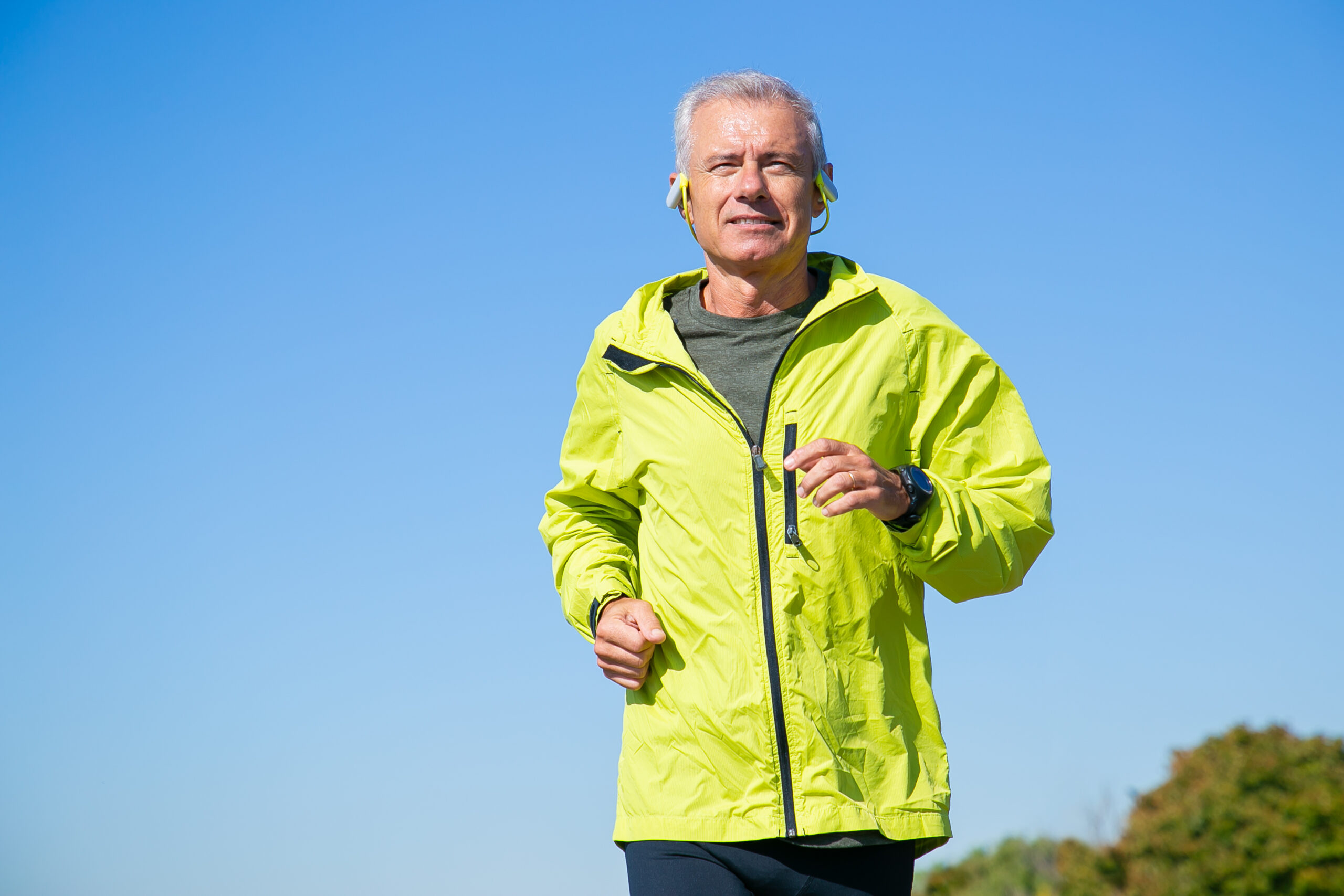 Happy excited senior man in wireless headphones jogging outside. Low angle, blue clear sky in background. Front view, copy space. Activity and age concept